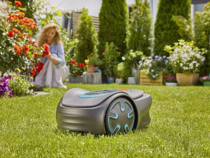 Een grijze robotmaaier voor een kleine tuin met lichtblauwe accenten op een groen gazon, met op de achtergrond een vrouw die rode rozen verzorgt.