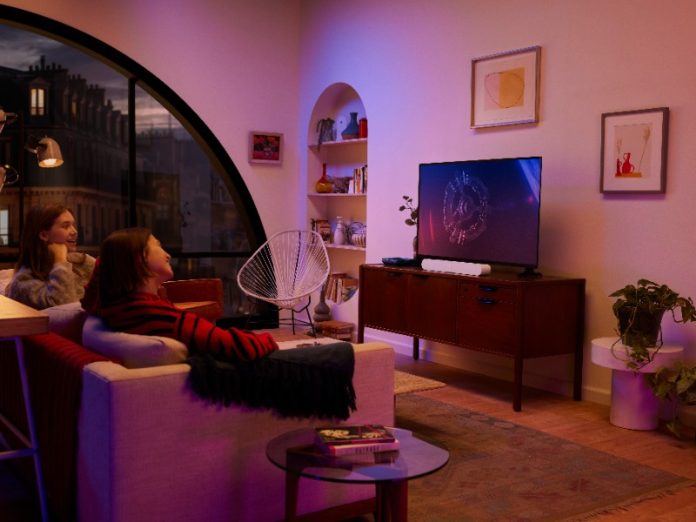 Twee vrouwen kijken televisie in een sfeervol verlichte woonkamer met paarse en roze accenten. Onder de tv staat een witte Sonos soundbar op een houten dressoir en door een groot boogvenster is een stad bij nacht zichtbaar.