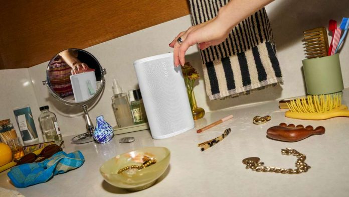 Een witte Sonos luidspreker op een badkamerplank tussen een gele haarborstel, een saliegroene tandenborstelhouder en diverse verzorgingsproducten voor een Sonos play review, terwijl een hand met natte vingers de bovenkant aanraakt.