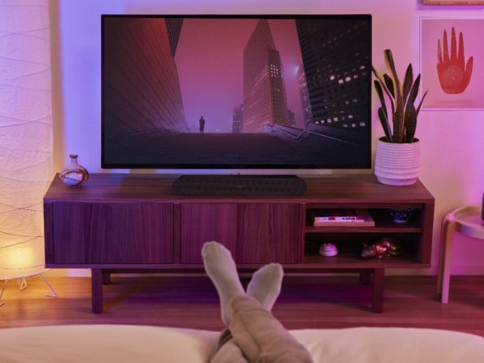 Zwarte Sonos soundbar op een houten tv-meubel onder een tv met rood-paars stadsgezicht, in een sfeervolle kamer met paarse belichting. Voeten in witte sokken op de voorgrond suggereren een ontspannen kijkervaring, relevant voor 'Sonos Soundbars vergeleken'.