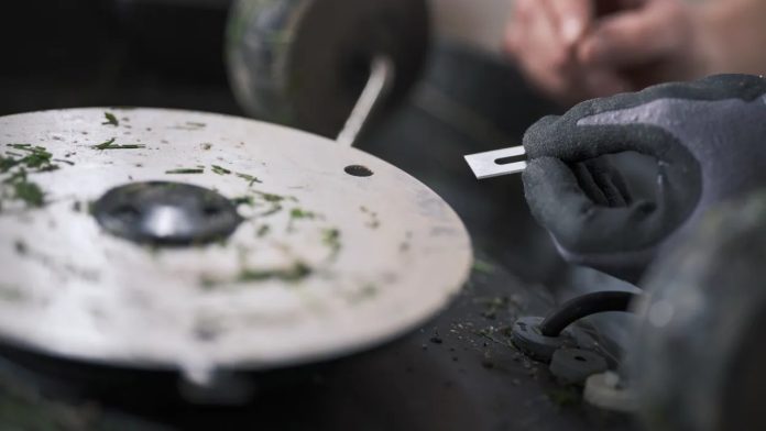 Een hand met een grijze werkhandschoen houdt een nieuw zilveren reservemesje bij de snijschijf van een robotmaaier om te tonen wanneer moet je de mesjes van je robotmaaier vervangen?