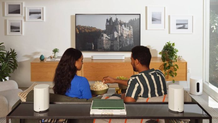 Een koppel kijkt tv in een moderne woonkamer met een surround-opstelling van 2x Sonos Era 100 of 1x Era 300, hier afgebeeld met twee witte Era 100-speakers op een zwarte sidetable achter de bank, een witte soundbar en een witte subwoofer.