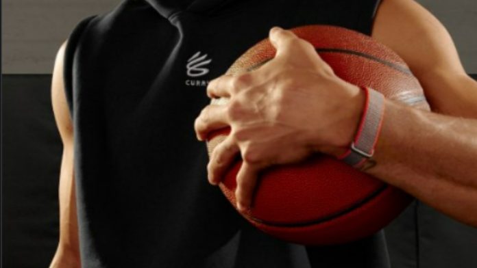 Stephen Curry in een zwarte mouwloze hoodie houdt een basketbal vast, met een grijze en rode polsband van de Fitbit Band leak om zijn pols.