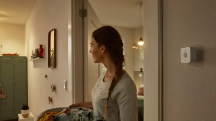 Een vrouw met een vlecht draagt een wasmand vol geel en blauw wasgoed door een sfeervol verlichte kamer langs een witte Philips Hue bewegingssensor aan de muur, een scenario waarbij de Hue bewegingssensor werkt niet in donker.