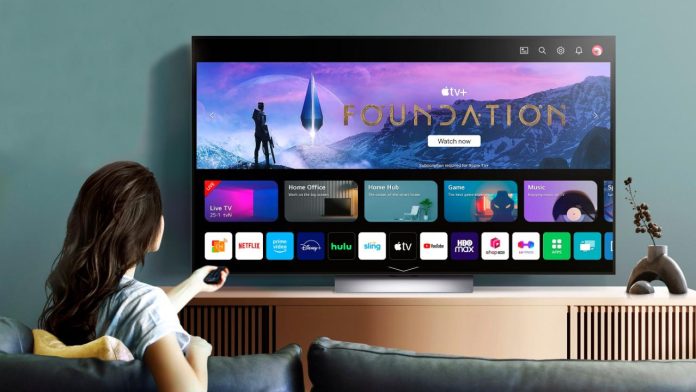 Een vrouw bedient een LG smart-tv met een kleurrijk menu vol streaming-apps vanaf een donkergrijze bank tegen een blauwgroene muur.