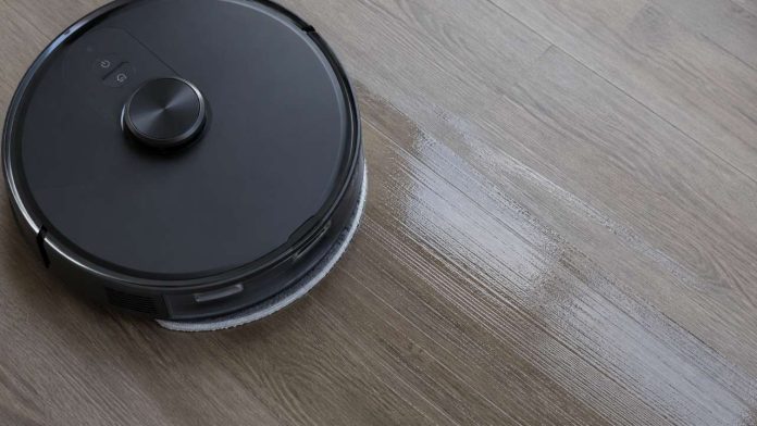 Een zwarte robotstofzuiger laat strepen achter op een lichtbruine houten vloer tijdens het dweilen.
