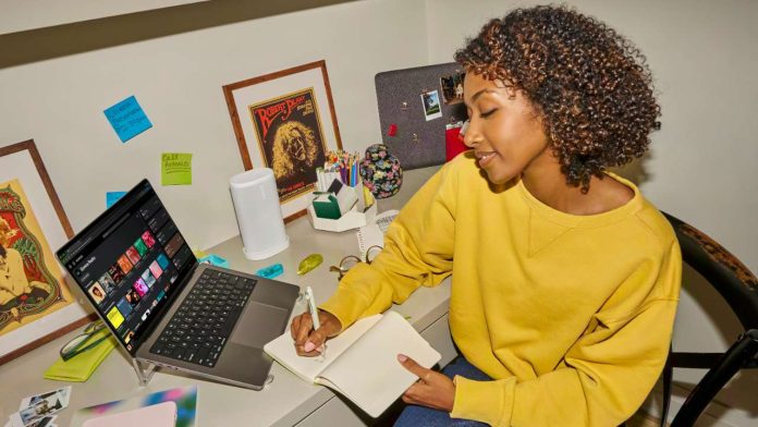 Een vrouw in een gele trui schrijft aan haar bureau naast een witte Sonos-speaker en een laptop met de Sonos-app voor het Sonos EQ instellen.