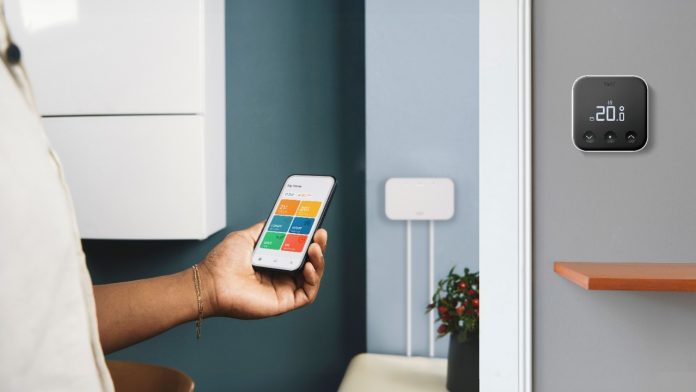 Een persoon houdt een smartphone vast met de kleurrijke tado-app geopend voor een donkerblauwe muur, met op de voorgrond een zwarte tado slimme thermostaat aan een grijze wand die 20,0 graden weergeeft.