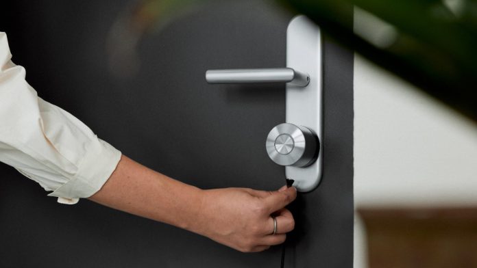 Een detailopname van een hand die een zwarte connector in een modern zilverkleurig slim deurslot op een antracietgrijze deur steekt, als illustratie bij de vraag: Kan Een Slim Deurslot Gehackt Worden?