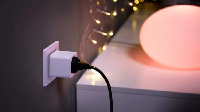 Een witte Philips Hue Smart Plug in een wandstopcontact met een zwarte stroomkabel, omgeven door sfeervolle, warm oranje verlichting.