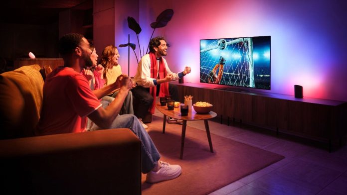 Vier vrienden juichen enthousiast in een woonkamer tijdens een live voetbalwedstrijd met Philips Hue Sports Live. De wand achter de televisie baadt in een sfeervolle, kleurrijke gloed van roze, paars en blauw licht dat de actie op het scherm volgt.