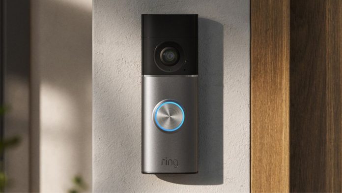 Zilveren en zwarte Ring Video Doorbell met een oplichtende blauwe ring gemonteerd op een witte muur naast een houten kozijn, ideaal om je Ring deurbel aan Alexa routines te koppelen.