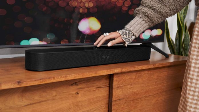 Een hand met een zilveren ring raakt een zwarte Sonos soundbar aan op een houten meubel voor een televisie, een handeling die past bij het Ring Deurbel Aan Alexa Routines Koppelen voor een slimme woonervaring.