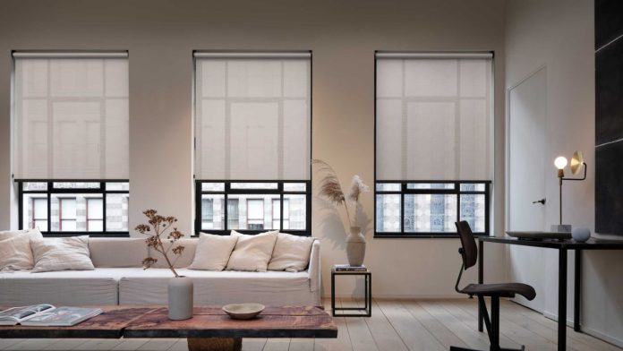 Drie lichtgrijze rolgordijnen in een modern, minimalistisch interieur laten zien hoe u uw slimme zonwering automatiseren kunt voor optimaal licht en comfort.