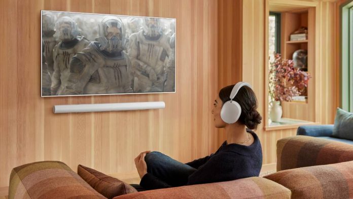 Een vrouw met een witte Sonos-koptelefoon en soundbar kijkt naar een film in een modern interieur met licht houten wanden, passend bij de Sonos AI toekomstplannen voor een naadloze audio-ervaring.