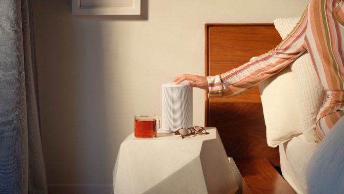 Een hand in een kleurrijke gestreepte pyjama raakt een witte Sonos-speaker aan op een wit geometrisch nachtkastje. Deze Sonos Wekker staat in een zonnige slaapkamer naast een houten bed met een glas thee en een bril.