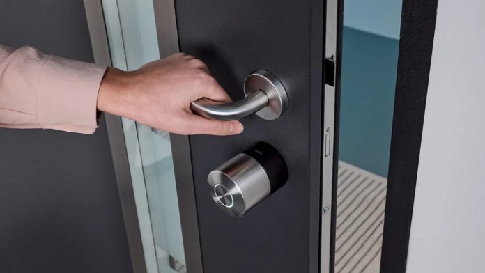 Een hand met een lichtroze mouw grijpt de zilveren kruk van een donkergrijze stroeve voordeur die voorzien is van een zilverkleurig slim deurslot.