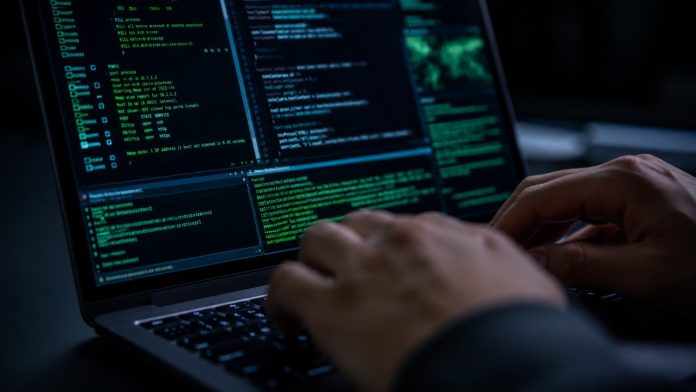 Iemand typt op een laptop met meerdere vensters groene en blauwe computercode op het scherm, wat een datalek hotels smart home symboliseert in een donkere omgeving.