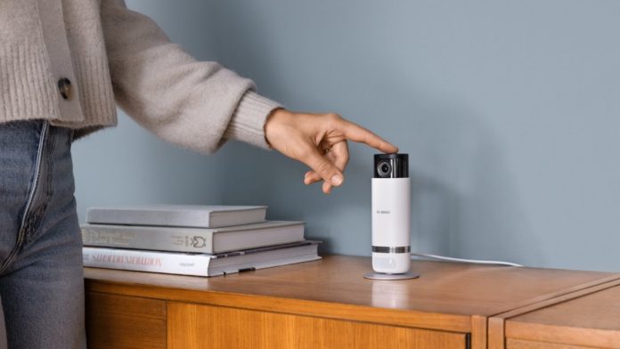 Een persoon in een lichtgrijs vest raakt een witte binnencamera op een houten dressoir aan tegen een blauwgrijze muur, een voorbeeld van de beste binnencamera's met privacy shutter.