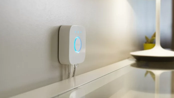 Een witte Philips Hue Bridge met oplichtende blauwe indicatielampjes is strak aan de muur bevestigd boven een wit bureau.