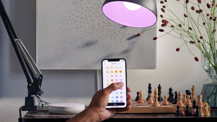 Een hand past via een smartphone de kleur aan van een IKEA DUBBELKISEL Matter-lamp naar een paarse tint boven een bureau met een schaakbord.