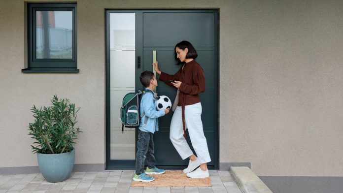 Een vrouw in een bruine top en witte broek opent een donkergroene voordeur met haar smartphone voor een jongen met een groene rugzak en voetbal, passend bij een vergelijking tussen Nuki vs SwitchBot.