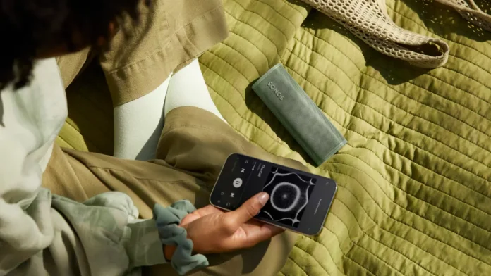 Een persoon bedient muziek via een smartphone bij een olijfgroene draagbare sonos luidspreker die op een groen gewatteerd kleed ligt.
