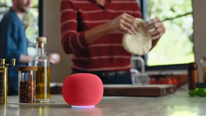 Een rode Google Home Speaker met Gemini AI staat op een aanrecht in de keuken terwijl een persoon op de achtergrond deeg bereidt.