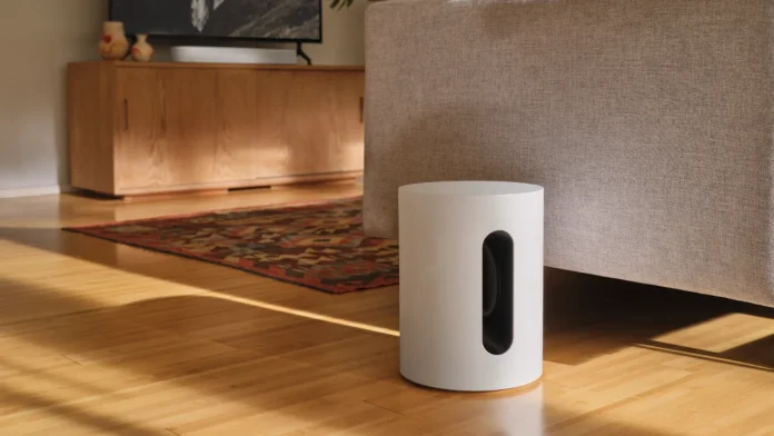De witte Sonos Sub Mini staat op een lichte houten vloer naast een grijze stoffen bank in een zonovergoten woonkamer.
