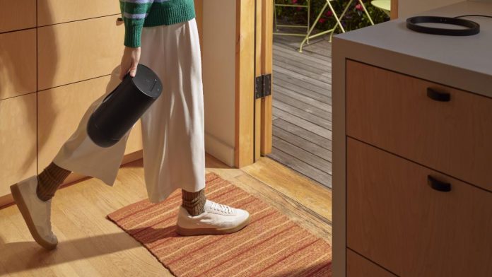 Een persoon in een groen en blauw gestreepte trui en wijde witte broek draagt een zwarte Sonos Move-speaker vanuit een lichte houten keuken naar een zonovergoten terras met een lichtgroene tuinset.