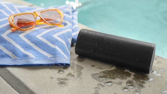Een zwarte Sonos-speaker bedekt met waterdruppels op een natte zwembadrand naast een blauw-witte handdoek en een oranje zonnebril.