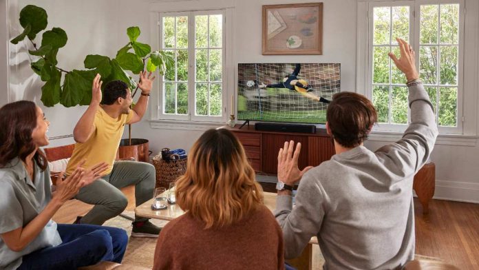 Vier enthousiaste vrienden juichen in een lichte woonkamer bij een Sonos WK 2026 opstelling. Terwijl een man in een geel shirt en een man in een grijze trui hun handen heffen naar de tv, is er een zwarte Sonos soundbar te zien onder het scherm op een houten dressoir, omgeven door witte muren en een grote groene kamerplant.