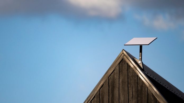Witte rechthoekige Starlink schotel gemonteerd op de nok van een donker houten dak tegen een helderblauwe lucht; een ideale oplossing voor wie een Starlink schotel huren overweegt.