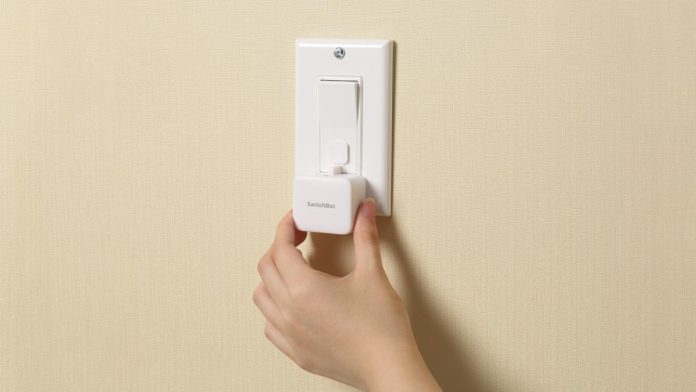 Een hand die een wit SwitchBot-apparaat op een lichtschakelaar op een beige muur monteert, in het kader van het bericht SwitchBot neemt Nanoleaf over.