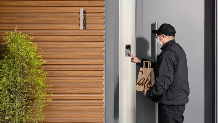 Een bezorger met een mondkapje drukt op de deurbel van een modern huis waar een videodeurbel oververhit hittegolf risico's kan lopen door direct zonlicht.