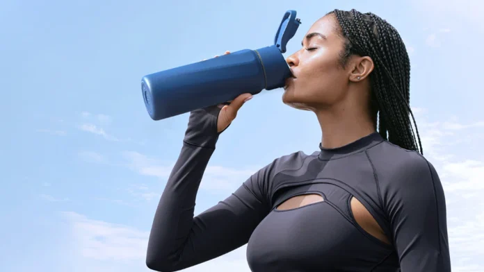 Een sportieve vrouw met vlechten drinkt verfrissend Water uit een grote blauwe fles tegen een heldere blauwe lucht.