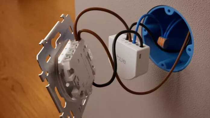 Een witte Philips Hue wandschakelaarmodule wordt met bruine en zwarte draden geïnstalleerd in een blauwe inbouwdoos achter een lichtschakelaar.