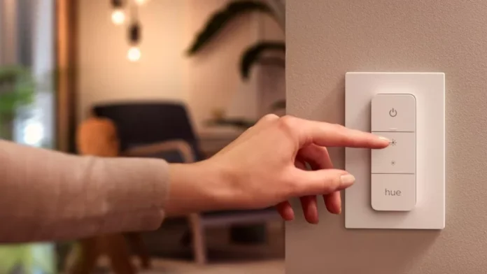 Een hand bedient een witte Hue dimschakelaar aan de muur om de verlichting in een sfeervolle woonkamer aan te passen.