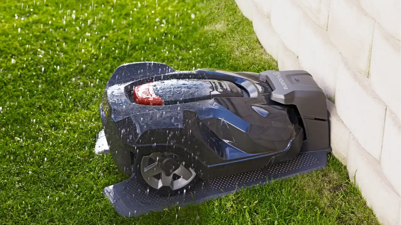 Een Husqvarna Automower 415X robotmaaier staat in zijn laadstation tegen een witte muur terwijl het regent op het groene gazon.
