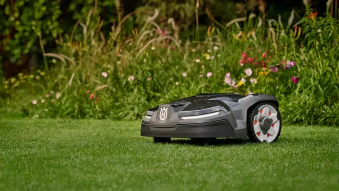 Een grijze Husqvarna Automower 415X robotmaaier maait een strak groen gazon langs een weelderige border vol kleurrijke bloemen.