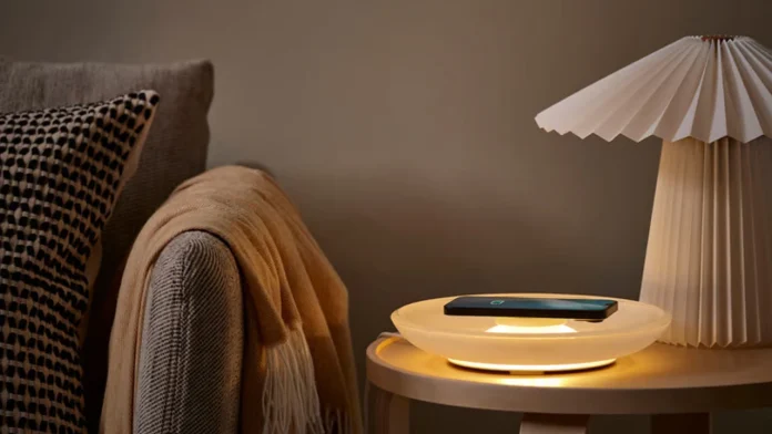 Een smartphone wordt draadloos opgeladen op de sfeervol verlichte IKEA VÄSTMÄRKE schaal die op een houten bijzettafeltje naast een gezellige bank staat.