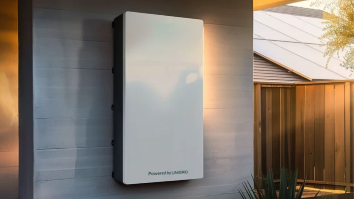 Een moderne witte natrium thuisbatterij van Unigrid hangt gemonteerd aan een grijze buitenmuur van een woning in het warme namiddaglicht.