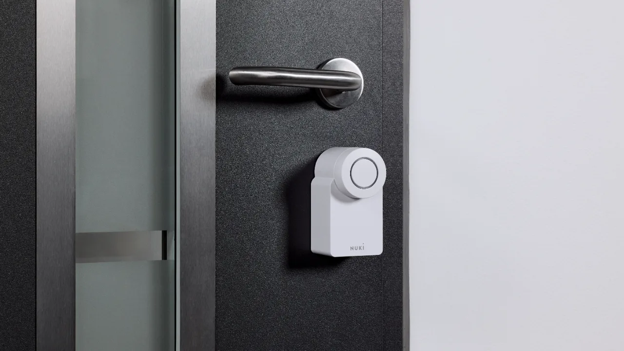 Een wit Nuki Smart Lock gemonteerd op een donkergrijze deur onder een metalen klink helpt je bij het Nuki Smart Lock vergelijken voor jouw slimme woning.
