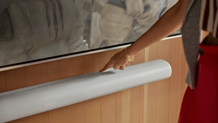 Een hand bedient de aanraakgevoelige knoppen van een witte Sonos soundbar die aan een stijlvolle houten wand onder de televisie is gemonteerd.