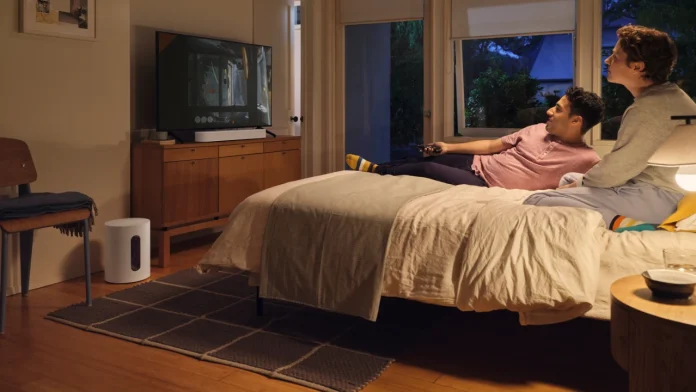 Een koppel kijkt ontspannen tv in een sfeervolle slaapkamer met een witte Sonos Sub appartement naast het houten dressoir.