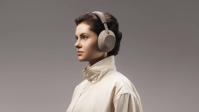 Een vrouw in een lichtgekleurde jas draagt de beige Sony WH-CH730N noise-cancelling hoofdtelefoon tegen een neutrale grijze achtergrond.