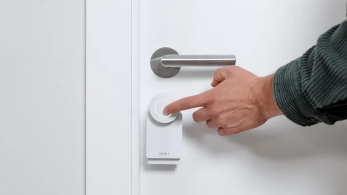 Een hand drukt op de knop van een wit Nuki slim deurslot op een witte deur.
