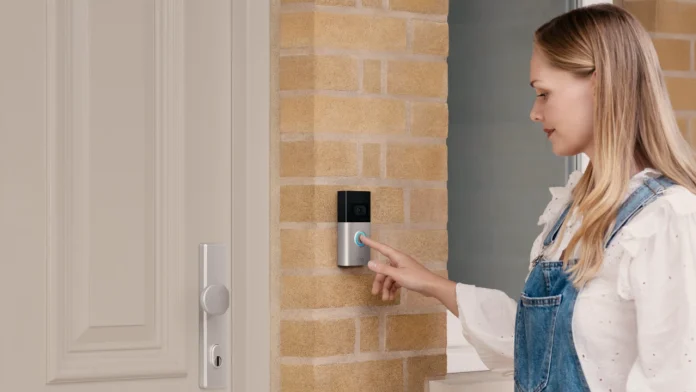 Een blonde vrouw drukt op de knop van een Ring videodeurbel die gemonteerd is op een bakstenen buitenmuur naast een witte voordeur.