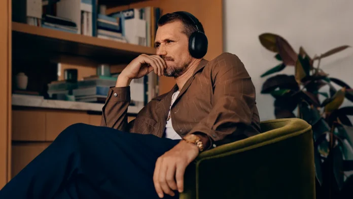 Peinzende man met zwarte Sonos koptelefoon in een groene fauteuil voor een boekenkast.