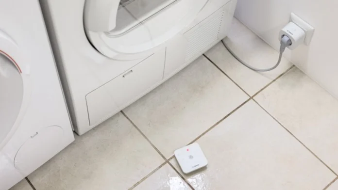 Een compacte witte WaterMelder signaleert een waterlekkage op de tegelvloer naast een witte wasmachine.